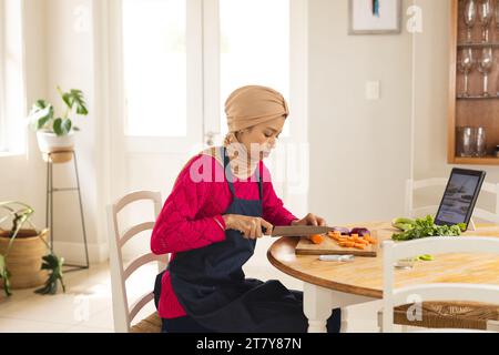 Birassische Frau in Hijab und Schürze, die Gemüse mit Tablette in der Küche zerkleinert Stockfoto