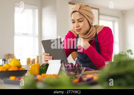 Glückliche birassische Frau in Hijab und Schürze mit Tablette in der Küche Stockfoto