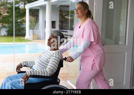 Glückliche kaukasische Krankenschwester und ältere afroamerikanische Patientin im Rollstuhl Stockfoto
