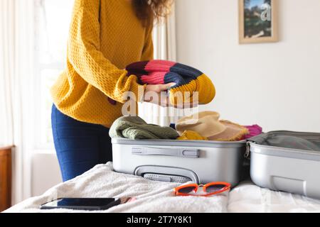 Mittelteil einer birassischen Frau, die Koffer auf dem Bett im sonnigen Schlafzimmer packt Stockfoto