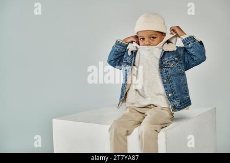 Niedlicher afroamerikanischer Junge in Winterkleidung und Mütze, der auf Betonwürfel sitzt und eine verstellbare Kapuze trägt Stockfoto