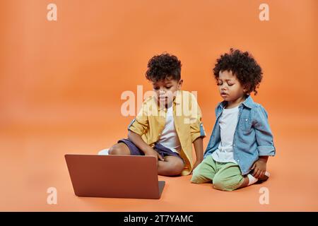 Niedliche afroamerikanische Geschwister in legeren Sommerbekleidung, die Filme auf einem Laptop anschauen, vor orangefarbenem Hintergrund Stockfoto