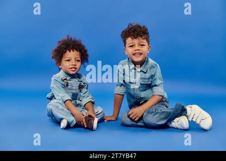 Niedliche afroamerikanische Geschwister in stilvoller Denim-Kleidung sitzen zusammen auf blauem Hintergrund Stockfoto