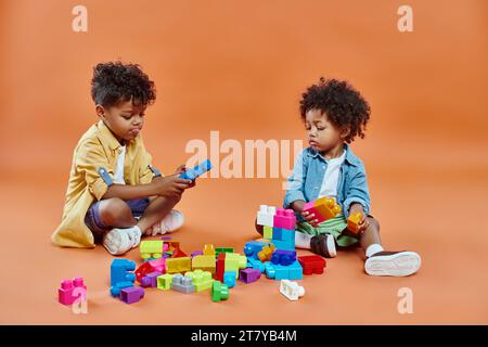 Niedlicher afroamerikanischer Geschwisterjunge in lässiger Kleidung, sitzend und spielend Bausteine auf Orange Stockfoto