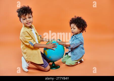 Glückliche afroamerikanische Geschwister in lässiger Kleidung berühren den Globus und sitzen vor orangefarbenem Hintergrund Stockfoto