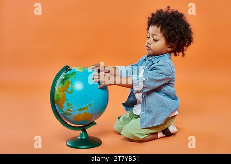 Glücklicher afroamerikanischer Kleinkind in legeren Kleidern, der den Globus berührt und vor orangefarbenem Hintergrund sitzt Stockfoto