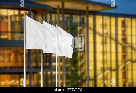 Weiße Firmenflaggen auf Fahnenmasten vor verschwommenem Bürogebäude mit Abendlicht. Leeres Flag-Modell zum Hinzufügen eines Logos, Symbols oder Schildes Stockfoto