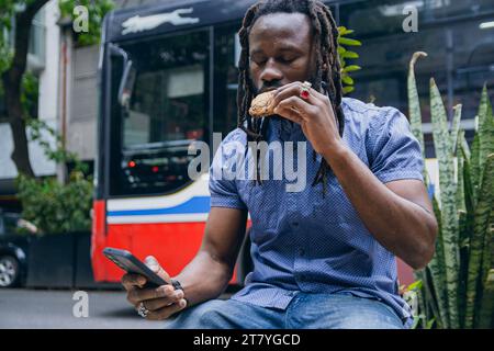 Junger schwarzer afrikanischer Mann mit Dreadlocks und Bart, sitzt mit dem Handy und überprüft das Telefon und isst draußen Kekse. Lifestyles-Konzept, Copy Spa Stockfoto