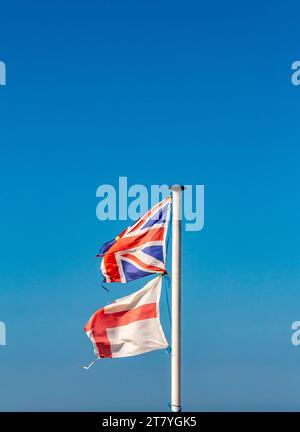 Zerrissene und zerrissene England- und Union Jack-Flaggen auf einem Fahnenmast mit blauem Himmel im Hintergrund. Stockfoto