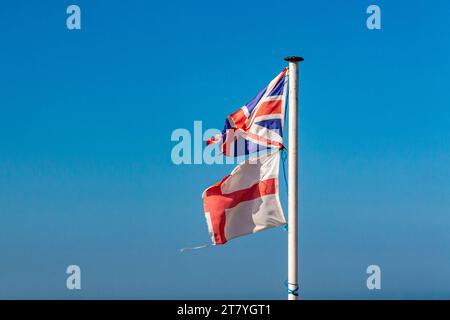 Zerrissene und zerrissene England- und Union Jack-Flaggen auf einem Fahnenmast mit blauem Himmel im Hintergrund. Stockfoto