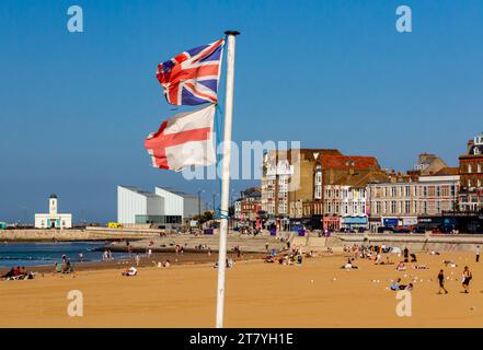 Sandstrand und Promenade in Margate, einem Badeort an der Küste von Kent im Südosten Englands, Großbritannien, mit zerrissenen englischen und britischen Flaggen an einem Pol. Stockfoto