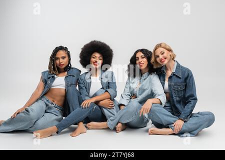 Fröhliche und barfuß multikulturelle Freundinnen in blauen Jeans, die vor grauem Hintergrund sitzen Stockfoto
