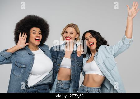 Aufregende multirassische Freundinnen in blauer Jeansbekleidung, die Hände auf grauer Streetmode winken Stockfoto