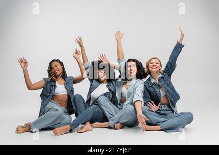 Aufgeregte und barfuß multirassische Freundinnen in Denim-Kleidung sitzen und Hände in Grau winken Stockfoto