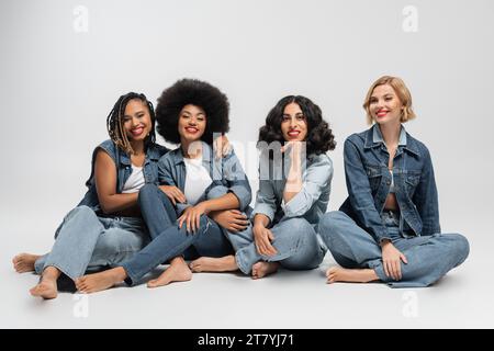 Fröhliche und barfuß multikulturelle weibliche Models in blauer Jeansbekleidung vor grauem Hintergrund Stockfoto