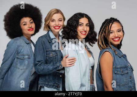 Fröhliche multikulturelle weibliche Models in lässiger Jeansbekleidung lächeln vor der Kamera auf grauer, vielfältiger Schönheit Stockfoto