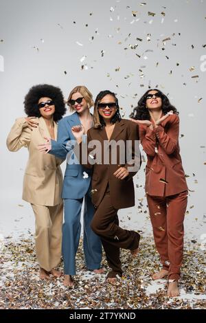 Fröhliche multirassische Freundinnen in Sonnenbrille und stylischen Anzügen, nahe glitzerndem Konfetti auf grauer Party Stockfoto