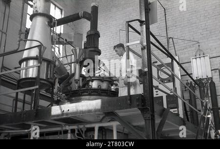 1960er Jahre, historisch, ein männlicher Agent, in schmutzigen Arbeitsanzügen, auf einer hohen Plattform stehend, an der Ausrüstung einer Lackierfabrik in England, Großbritannien. Stockfoto