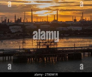 Silhouetten der Ölraffinerie und der Lagertanks während eines lebhaften Sonnenuntergangs, der sich auf einer ruhigen Flussoberfläche in Southampton UK spiegelt Stockfoto