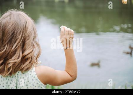 Unerkennbares Mädchen in hellem Kleid steht mit dem Rücken und füttert Enten in der Nähe des Teichs in der Natur. Stockfoto