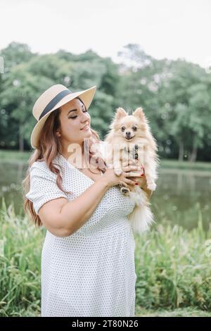 Schöne junge Frau im Sommerkleid und Hut steht mit kleinen pommern in den Armen in der Natur. Stockfoto