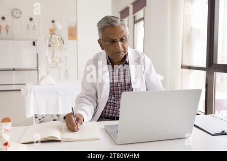Reifer indischer Arzt im Uniformmantel, der am Laptop arbeitet Stockfoto