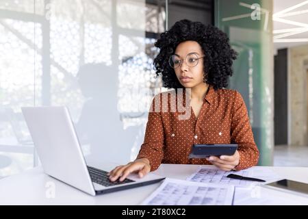 Ernsthaft denkende Mitarbeiterin Finanzierin, die mit Dokumenten und Papieren am Arbeitsplatz arbeitet, Geschäftsfrau, die Berichte prüft, Taschenrechner und Laptop in Papierkram verwendet. Stockfoto