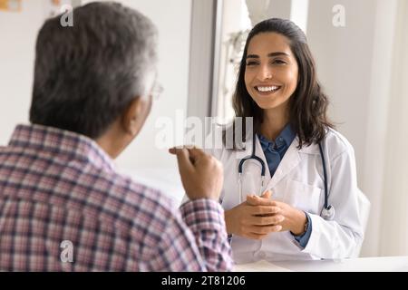 Fröhliche junge lateinische geriatrische Ärztin, die dem sprechenden Mann zuhört Stockfoto