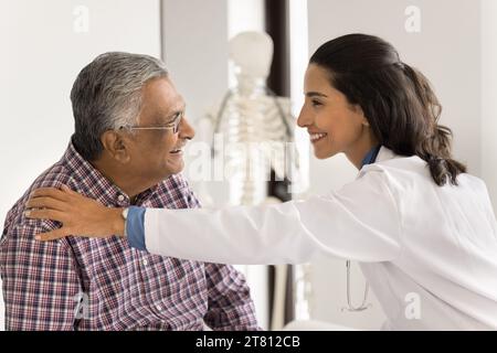 Fröhliche junge Ärztin, die die Schulter eines älteren indischen Patienten berührt Stockfoto