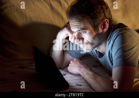 Junger gutaussehender Mann, der nachts im Bett liegt und etwas auf einem Tablet sieht. Kopierbereich für Text. Stockfoto