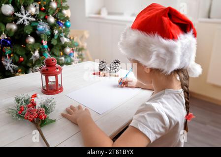 Das Mädchen schreibt dem Weihnachtsmann zu Hause einen Brief mit Weihnachtsmütze. Baum- und festliche Dekoration im Hintergrund. Herzerwärmende Urlaubsszene Stockfoto