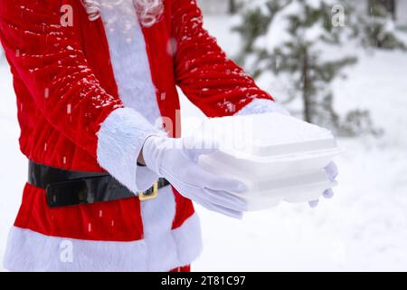 Lebensmittelliefercontainer in den Händen des Weihnachtsmanns im Freien im Schnee. Weihnachtsabend-Angebot. Gebrauchsfertige heiße Bestellung, Einweg-Kunststoffbox. Neues y Stockfoto