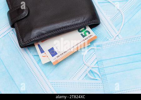 Schwarze Herren Brieftasche mit Euro Geld innen auf den blauen Einweg-medizinischen Gesichtsmasken. Handel mit medizinischen Masken. Unternehmen während einer Pandemie oder Epidemie Stockfoto