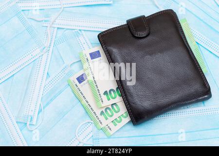 Schwarze Herren Brieftasche mit Euro Geld innen auf den blauen Einweg-medizinischen Gesichtsmasken. Handel mit medizinischen Masken. Unternehmen während einer Pandemie oder Epidemie Stockfoto