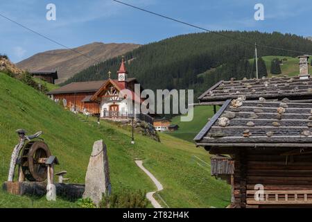 Rosenkranzkapelle, Almdorf Gemais, Außenansicht, Eingang, Vorderseite, Hanglage, Turmuhr, Brunnen, alpine Bergwelt, Nadelbäume Stockfoto