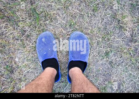 Haarige Männerbeine in schwarzen Socken, in alten blauen Hausschuhen mit Löchern, stehen auf einem halbgetrockneten Rasen im Innenhof des Hauses Stockfoto