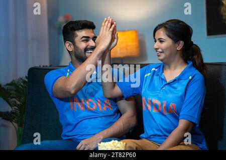 Begeisterte Happy Couples Indian Team Trikot feiert Sieg oder Sieg beim Fernsehen oder Fernsehen zu Hause - Konzept der Unterhaltung, Sportfans Stockfoto