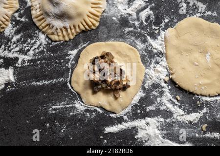 Herstellung glutenfreier Teigtaschen mit Pilzen und Zwiebeln. Dunkelgraue Arbeitsplatte mit Mehl bestreut. Hausgemacht. Stockfoto