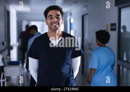 Porträt eines glücklichen, birassischen männlichen Arztes, der im Krankenhauskorridor steht Stockfoto