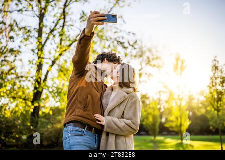 Porträt eines glücklichen Liebespaares im Park bei Sonnenuntergang. Sie küssen und machen Selfie. Stockfoto