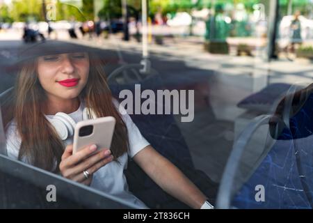 Foto durch das Glas des Busfensters, Passagierin, die ihr Smartphone benutzt, während sie im Shuttlebus sitzt. Stockfoto