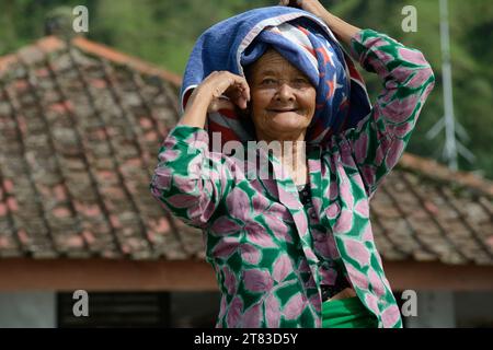 Eine balinesische ältere Frau, deren Gesicht mit der Weisheit der Jahre geätzt ist, steht stolz in traditioneller Kleidung vor dem Hintergrund ihres Dorfhauses Stockfoto