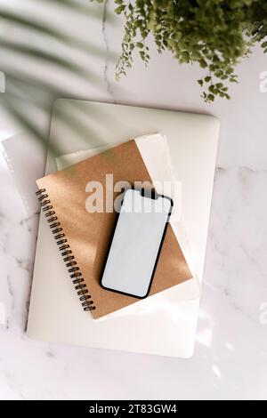 Mobiltelefon mit leerem Pfadbildschirm und Mockup auf verschiedenen Notizblöcken und geschlossenem Laptop bei Sonnenlicht mit Schatten. Ästhetisches Social Media Lifestyle Konzept. T Stockfoto