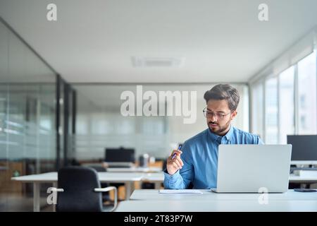 Beschäftigter junger lateinamerikanischer Geschäftsmann, der im Büro arbeitet und Dokumente mit Laptop überprüft. Stockfoto