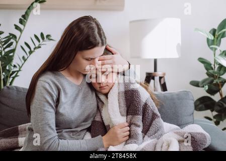 Liebevolle junge Mutter umarmt kranke, verärgerte, kleine Tochter bedeckte Decke, sitzt zusammen auf der Couch zu Hause, fürsorgliche Mutter tröstet trauriges, krankes Kind, expressi Stockfoto