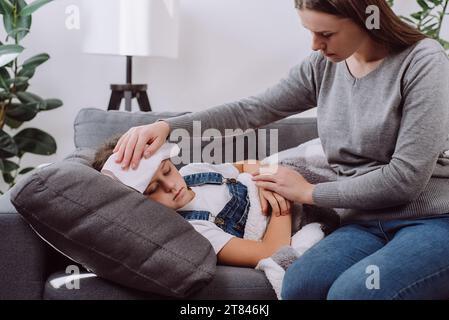 Die fürsorgliche junge Mutter kümmert sich um die kleine kranke Tochter. Das kranke Kind hat hohes Fieber, liegt auf der Couch unter der Decke. Alleinerziehende kaukasische Mutter kümmert sich um Kranke Stockfoto