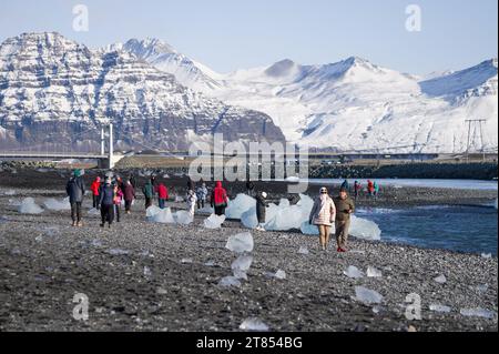 Touristen, die entlang der Küste von Breiðamerkursandur, oder Diamond Beach, an der Lagune von Jokulsarlon spazieren, mit Eisbergen, die an der Küste in Island angespült werden Stockfoto
