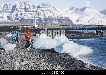 Touristen, die entlang der Küste von Breiðamerkursandur, oder Diamond Beach, an der Lagune von Jokulsarlon spazieren, mit Eisbergen, die an der Küste in Island angespült werden Stockfoto