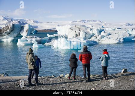 Touristen stehen am Ufer der Gletscherlagune Jökulsárlón mit Eisbergen, die in der Lagune in Island schwimmen Stockfoto