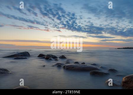 Romantische Atmosphäre bei friedlichem Sonnenuntergang am Meer. Große Felsbrocken, die aus glattem, welligem Meer ragen. Gelber Streifen am Horizont und winzige Wolken Stockfoto